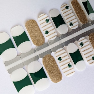 Könnte beinhalten: Nagelaufkleber mit St. Patrick's Day-Motiv. Designs umfassen goldfarbenen Glitzer, Weiß mit goldenen Streifen und Kleeblättern sowie Weiß mit grünen Akzenten. Die Aufkleber befinden sich auf einer transparenten Folie.