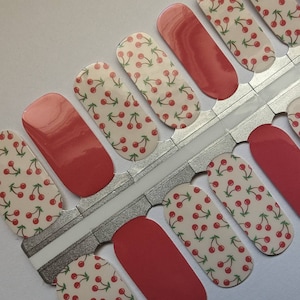 May include: Nail polish wraps with a cherry pattern on a white background. The wraps are on a silver backing sheet with a red accent strip.