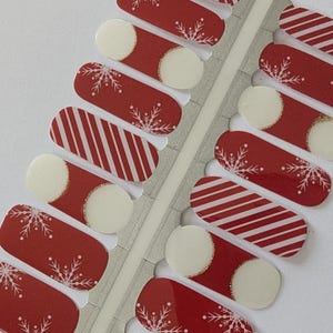 Op de afbeelding: Rode en witte nagelstrips met sneeuwvlok- en streepontwerpen. Elke strip heeft een afgerond wit uiteinde met een gouden glitteraccent. De strips zijn gerangschikt op een zilveren achtergrond, perfect voor een feestelijke manicure.