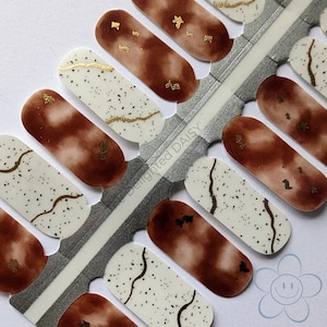 May include: Nail polish wraps with a brown and white watercolor design and gold accents. The wraps are arranged on a silver sheet with the text "delighted DAISY" printed on it.