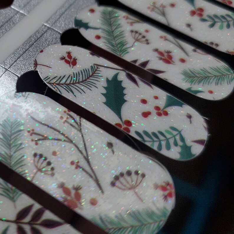 May include: Close-up of nail art strips with a winter holiday theme. The strips feature a white background with glitter and various designs, including holly, berries, and pine branches. The designs are in shades of green, red, and brown.