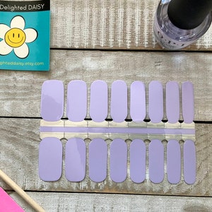 以下が含まれることがあります： ラベンダー色のネイルポリッシュラップのシートで、20枚の個別のラップが付属しています。ラップは2列10枚ずつに配置されています。シートは、ピンクのネイルファイルと木製のキューティクルプッシャーのある木製の表面の上にあります。