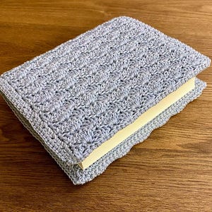 Op de afbeelding: Een lichtgrijze rechthoekige boekomslag van gehaakt garen met een herhalend schelpensteekpatroon. De hoes zit strak om een boek met cr&egrave;mekleurige pagina's en toont een gestructureerd, handgemaakt textielontwerp.
