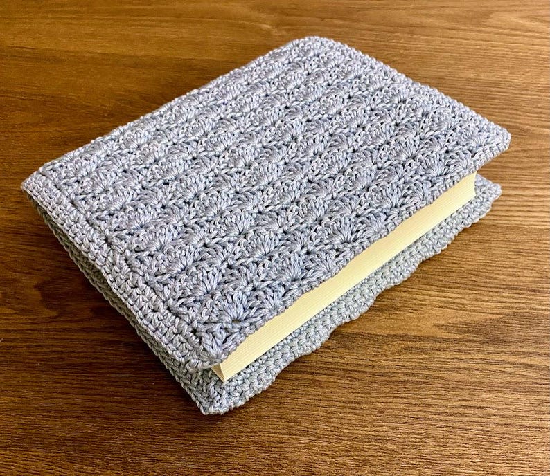 Op de afbeelding: Een lichtgrijze rechthoekige boekomslag van gehaakt garen met een herhalend schelpensteekpatroon. De hoes zit strak om een boek met cr&egrave;mekleurige pagina's en toont een gestructureerd, handgemaakt textielontwerp.