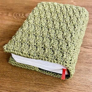 Op de afbeelding: Een rechthoekig boek in een saliegroene gehaakte hoes met een gestructureerd schelpenpatroon. De witte pagina's van het boek zijn aan de zijkant zichtbaar en een dun rood lint als bladwijzer steekt uit de onderkant van de hoes.