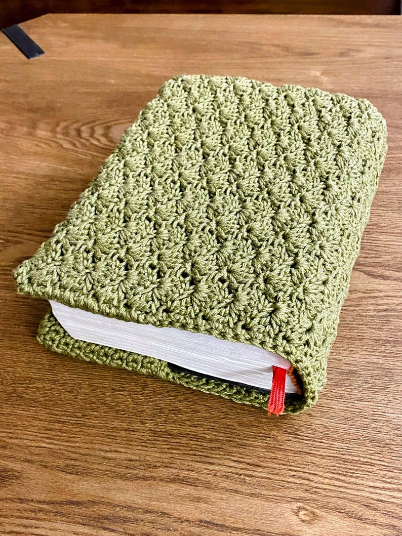 Op de afbeelding: Een rechthoekig boek in een saliegroene gehaakte hoes met een gestructureerd schelpenpatroon. De witte pagina's van het boek zijn aan de zijkant zichtbaar en een dun rood lint als bladwijzer steekt uit de onderkant van de hoes.