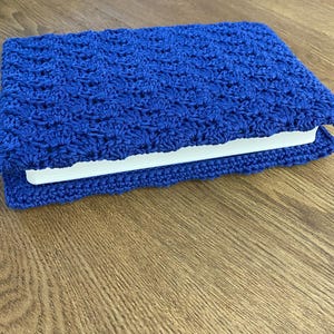 Op de afbeelding: Een rechthoekige boekomslag gemaakt van felblauw garen, met een gestructureerd, herhalend haakpatroon in schelpvorm. De omslag is om een wit boek gewikkeld, waarbij de randen van het haakwerk iets voorbij de rug van het boek uitsteken.