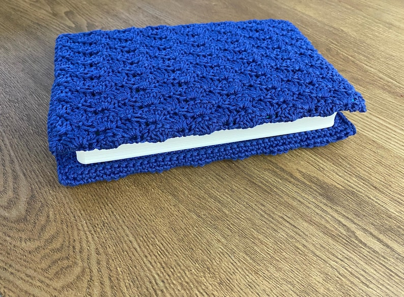 Op de afbeelding: Een rechthoekige boekomslag gemaakt van felblauw garen, met een gestructureerd, herhalend haakpatroon in schelpvorm. De omslag is om een wit boek gewikkeld, waarbij de randen van het haakwerk iets voorbij de rug van het boek uitsteken.