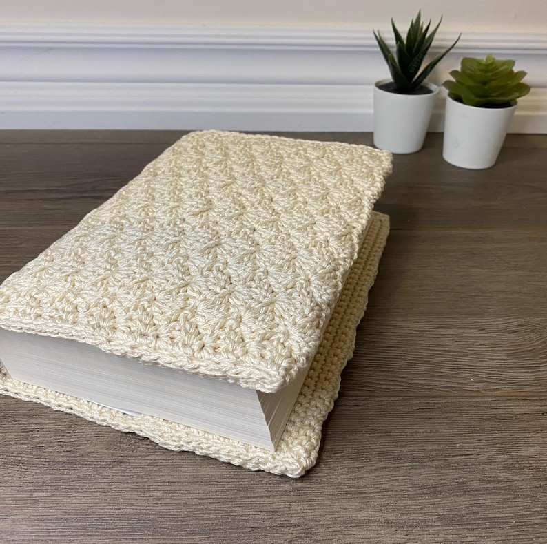 Op de afbeelding: Een rechthoekig boek in een cr&egrave;mekleurige, gehaakte hoes met textuur. Het garenwerk heeft een herhalend, ingewikkeld steekpatroon dat de voor-, achterkant en de rug van het boek volledig omsluit, wat zorgt voor een zachte, tastbare buitenkant.
