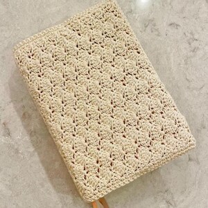 Op de afbeelding: Een rechthoekige boekomslag met een cr&egrave;mekleurig haakpatroon in een herhalend schelpsteekontwerp. De textiele omslag heeft een gestructureerd oppervlak en een klein bruin leren lipje zichtbaar aan de onderkant van het rechthoekige object.