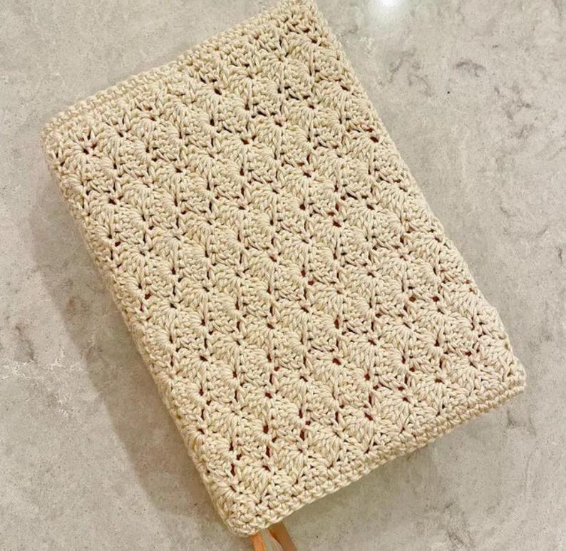 Op de afbeelding: Een rechthoekige boekomslag met een cr&egrave;mekleurig haakpatroon in een herhalend schelpsteekontwerp. De textiele omslag heeft een gestructureerd oppervlak en een klein bruin leren lipje zichtbaar aan de onderkant van het rechthoekige object.