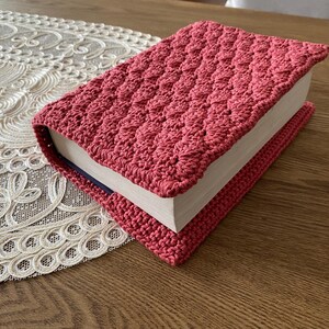 Op de afbeelding: Een rechthoekig boek in een gehaakte hoes met een textuur in koraalroze. Het haakwerk heeft een herhalend, schelpachtig steekpatroon dat om de voorzijde, achterzijde en de rug van het boek heen loopt, waarbij de witte pagina's zichtbaar zijn.