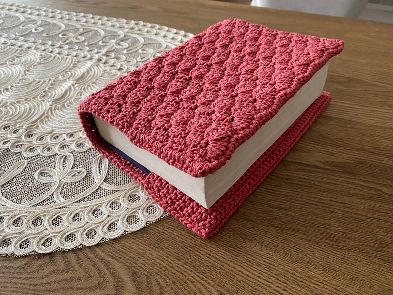 Op de afbeelding: Een rechthoekig boek in een gehaakte hoes met een textuur in koraalroze. Het haakwerk heeft een herhalend, schelpachtig steekpatroon dat om de voorzijde, achterzijde en de rug van het boek heen loopt, waarbij de witte pagina's zichtbaar zijn.