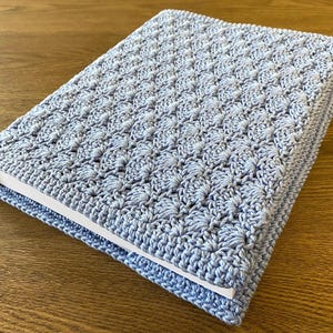 Op de afbeelding: Een rechthoekige boekomslag van lichtblauw garen, voorzien van een gestructureerd, herhalend schelpensteekpatroon. De gehaakte hoes zit strak om een boek met witte pagina's en vertoont een consistente, dichte en decoratieve vezelstructuur.