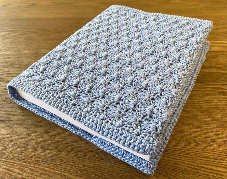 Op de afbeelding: Een rechthoekige boekomslag van lichtblauw garen, voorzien van een gestructureerd, herhalend schelpensteekpatroon. De gehaakte hoes zit strak om een boek met witte pagina's en vertoont een consistente, dichte en decoratieve vezelstructuur.