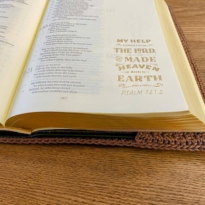 Op de afbeelding: Een open boek met cr&egrave;mekleurige pagina's en goudkleurige tekst die luidt: My help comes from the Lord, who made heaven and earth, Psalm 121:2. Het boek zit in een bruine, gehaakte hoes met een zichtbaar en gestructureerd steekpatroon.