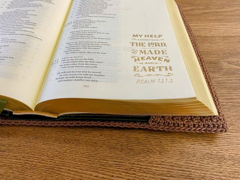Op de afbeelding: Een open boek met cr&egrave;mekleurige pagina's en goudkleurige tekst die luidt: My help comes from the Lord, who made heaven and earth, Psalm 121:2. Het boek zit in een bruine, gehaakte hoes met een zichtbaar en gestructureerd steekpatroon.