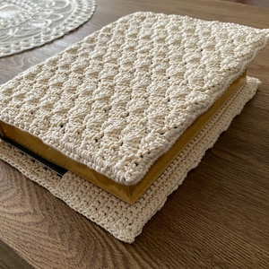 Op de afbeelding: Een rechthoekige boekomslag gemaakt van cr&egrave;mekleurig garen met een gestructureerd, herhalend haakpatroon. De omslag heeft een gevouwen flapontwerp dat om de randen van een boek met een goudkleurige rug en een donkere kaft heen valt.