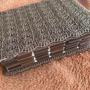 Op de afbeelding: Een boek met een getextureerde, taupekleurige gehaakte kaft met een herhalend schelppatroon. Het boek bevat meerdere tabbladen aan de zijkant, met zichtbare tekstlabels waaronder Joshua, Deuteronomy en Numbers.