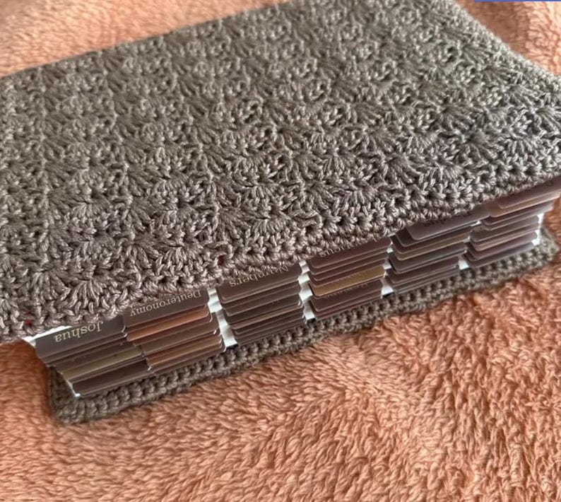 Op de afbeelding: Een boek met een getextureerde, taupekleurige gehaakte kaft met een herhalend schelppatroon. Het boek bevat meerdere tabbladen aan de zijkant, met zichtbare tekstlabels waaronder Joshua, Deuteronomy en Numbers.