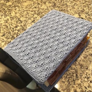 Op de afbeelding: Een lichtblauwe gehaakte boekomslag met een herhalend schelpenpatroon. De omslag zit om een boek heen met meerdere bruine tabbladen aan de zijkant, wat wijst op een gecategoriseerd systeem voor het ordenen van de pagina's in het boek.
