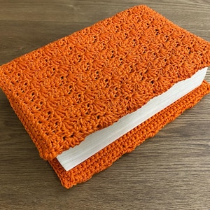 Op de afbeelding: Een rechthoekig boek in een feloranje gehaakte hoes met een herhalend stervormig steekpatroon. De hoes omsluit de voor- en achterkant van het boek, waardoor de witte pagina's zichtbaar zijn langs de zijkant van het gesloten boekdeel.