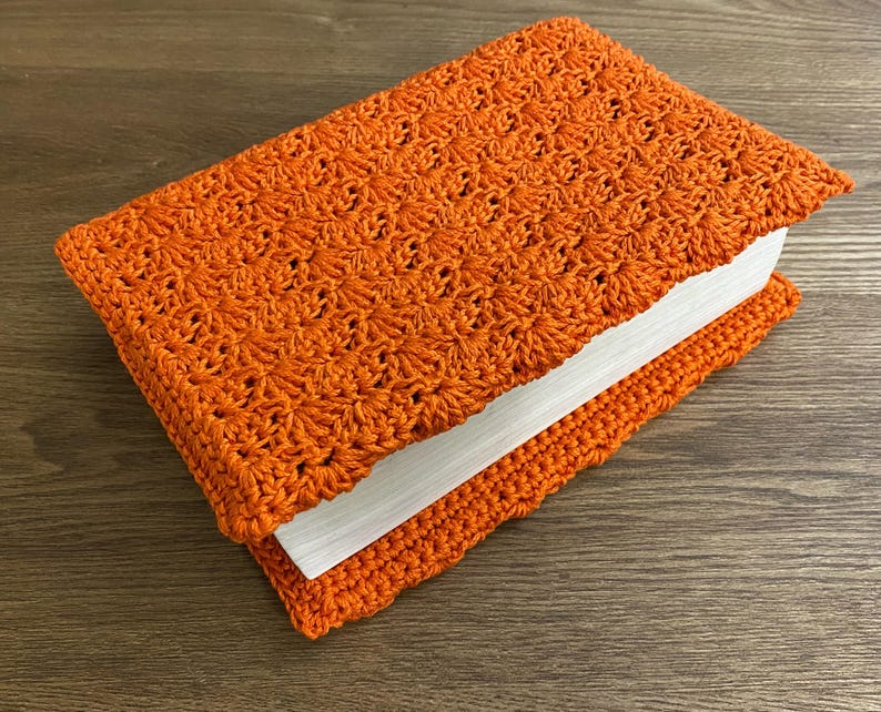 Op de afbeelding: Een rechthoekig boek in een feloranje gehaakte hoes met een herhalend stervormig steekpatroon. De hoes omsluit de voor- en achterkant van het boek, waardoor de witte pagina's zichtbaar zijn langs de zijkant van het gesloten boekdeel.