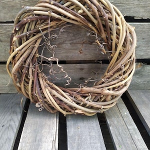 May include: A circular wreath made of intertwined, dried vines in shades of brown and tan. The wreath has a rustic, natural appearance, with some delicate, dried tendrils visible. The wreath is displayed on a weathered wooden surface.