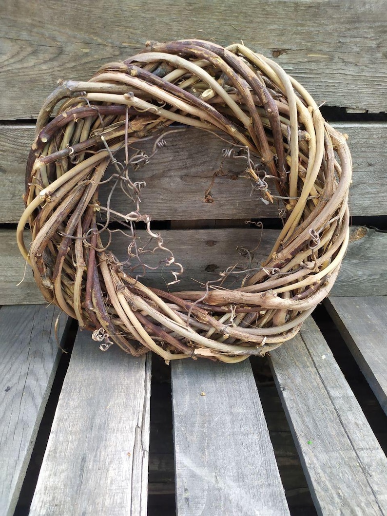 May include: A circular wreath made of intertwined, dried vines in shades of brown and tan. The wreath has a rustic, natural appearance, with some delicate, dried tendrils visible. The wreath is displayed on a weathered wooden surface.