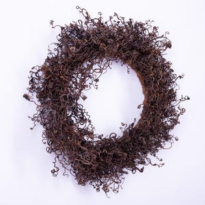 May include: A circular wreath composed of dried, brown, curly plant matter. The wreath is set against a plain white backdrop. The wreath's texture is dense and intricate, with numerous small curls and tendrils.