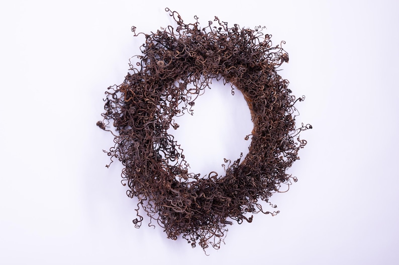 May include: A circular wreath composed of dried, brown, curly plant matter. The wreath is set against a plain white backdrop. The wreath's texture is dense and intricate, with numerous small curls and tendrils.