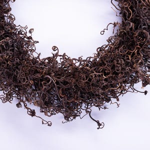 May include: A close-up of a decorative wreath made of dried, curly, brown plant material. The wreath is arranged in a circular shape against a white background. The texture of the dried plant material is visible.