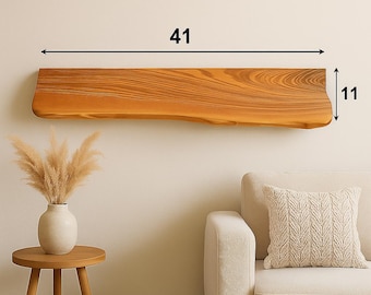 Pode incluir: Uma prateleira flutuante de madeira com borda natural, medindo 104 cm de comprimento e 28 cm de largura. A prateleira é de cor castanho claro quente com grão de madeira visível. É montada em uma parede de cor neutra acima de um sofá. A prateleira faz parte de um tema de decoração de casa.