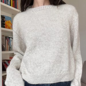 Könnte beinhalten: Ein hellgrauer Strickpullover mit Rundhalsausschnitt und geripptem Saum. Der Pullover wird mit einer dunkelblauen Jeanshose kombiniert. Das Outfit ist schlicht und lässig, geeignet für den Alltag.