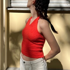 Könnte beinhalten: Ein leuchtend rotes Haltertop mit V-Ausschnitt. Das Oberteil wird mit einer weißen Hose kombiniert. Das Foto wurde im Freien aufgenommen, mit Sonnenlicht, das Schatten auf eine gelbe Wand und ein Fenster mit geschlossenen Jalousien wirft.