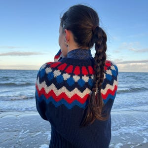 Könnte beinhalten: Ein marineblauer Pullover mit einem geometrischen Muster in Rot, Weiß und Blau. Der Pullover hat einen gerippten Saum und Bündchen. Die Person steht an einem Strand mit dem Meer im Hintergrund.