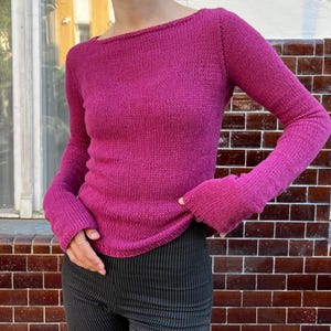 Op de afbeelding: Een magenta gebreide trui met lange mouwen en een boothals. De mouwen vallen wijd uit bij de polsen. De trui wordt gecombineerd met een zwart-wit gestreepte broek. De achtergrond is een bakstenen muur.