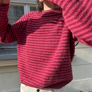 Könnte beinhalten: Ein rot-dunkelrosa gestreifter Pullover mit langen Ärmeln. Der Pullover ist aus Strickmaterial und hat einen runden Ausschnitt. Die Person trägt cremefarbene Hosen.