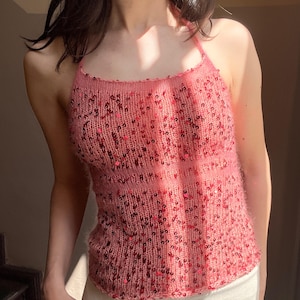 May include: A pink halter top with a knitted texture and embellished with sequins. The top has a scoop neckline and a fitted silhouette. The color is a soft, light pink with darker pink sequins scattered throughout.