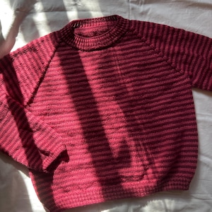 Puede incluir: Un su&eacute;ter a rayas burdeos y rosa oscuro con cuello redondo y mangas largas. La prenda de punto est&aacute; hecha de un material suave y es adecuada para climas m&aacute;s fr&iacute;os. El su&eacute;ter se muestra sobre una superficie blanca.