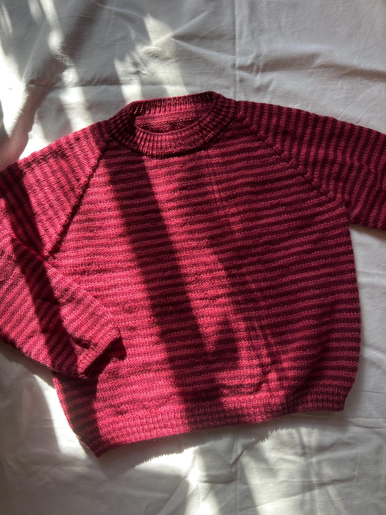 Puede incluir: Un su&eacute;ter a rayas burdeos y rosa oscuro con cuello redondo y mangas largas. La prenda de punto est&aacute; hecha de un material suave y es adecuada para climas m&aacute;s fr&iacute;os. El su&eacute;ter se muestra sobre una superficie blanca.