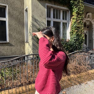 Puede incluir: Un su&eacute;ter burdeos con rayas horizontales y pantalones color crema. La persona est&aacute; de pie frente a un edificio con hiedra. Una valla de metal negro y una pared de ladrillo est&aacute;n en primer plano. El su&eacute;ter tiene rayas horizontales.