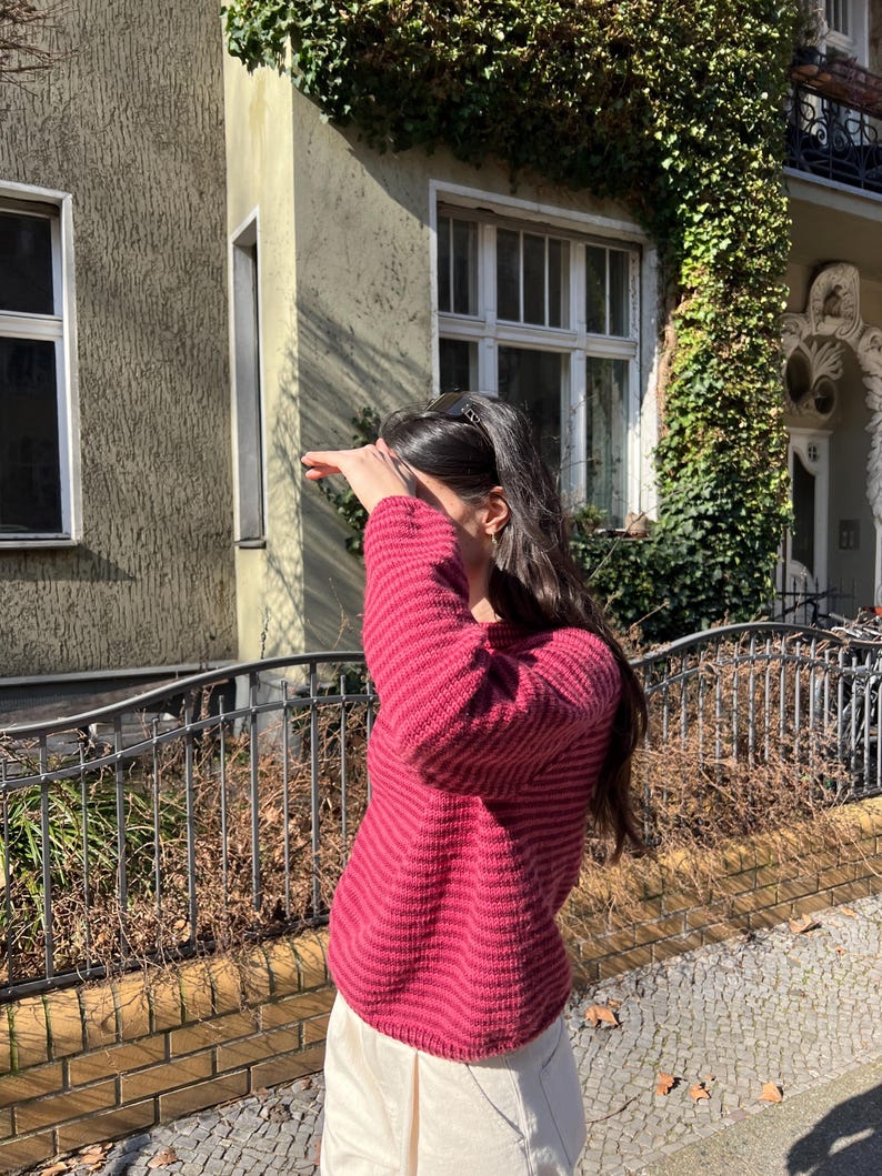 Puede incluir: Un su&eacute;ter burdeos con rayas horizontales y pantalones color crema. La persona est&aacute; de pie frente a un edificio con hiedra. Una valla de metal negro y una pared de ladrillo est&aacute;n en primer plano. El su&eacute;ter tiene rayas horizontales.