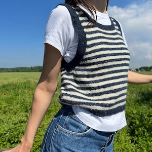 May include: A navy and cream striped knit vest over a white t-shirt and blue jeans. The vest has a rounded neckline and wide straps. The person is standing outside on a sunny day with a blue sky and green field in the background.