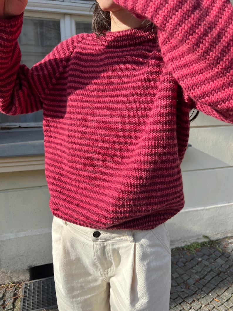 Puede incluir: Un su&eacute;ter a rayas rojas y rosa oscuro con mangas largas. El su&eacute;ter es de punto y tiene cuello redondo. La persona lleva pantalones color crema.