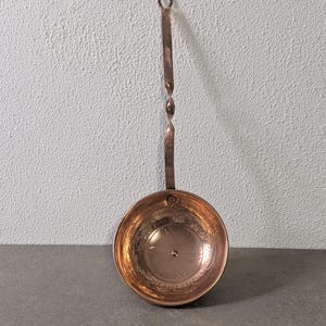 Könnte beinhalten: Eine Schöpfkelle aus poliertem Kupfer mit einer runden Schale, die mit geprägten Blumenmustern verziert ist, und einem langen, schlanken Griff, der einen dekorativen gedrehten Abschnitt sowie eine Öse zum Aufhängen am oberen Ende besitzt.