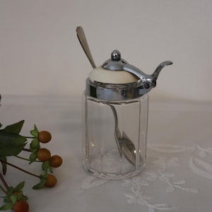 Puede incluir: Un azucarero de cristal vintage con tapa y asa de metal plateado. La tapa tiene un inserto color crema y una cucharita en su interior. El tarro de cristal transparente tiene un diseño facetado.