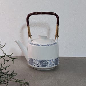 Könnte beinhalten: Weiße Keramik-Teekanne mit blauem Blumenmuster am Boden und am Rand. Sie hat einen braunen, gewebten Griff und einen passenden Deckel. Die Teekanne ist zum Aufbrühen von Tee bestimmt.