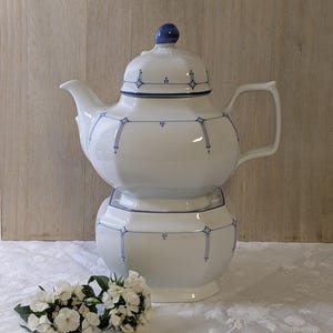 Puede incluir: Juego de tetera y azucarero de porcelana blanca con adornos azules decorativos. La tetera tiene una tapa a juego con un pomo azul. El juego se exhibe sobre un mantel blanco con un arreglo floral.