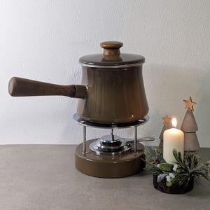 Può includere: Una fonduta marrone con manico e coperchio in legno è su un supporto cromato. Una candela accesa e alberi di Natale decorativi in legno sono a destra. La pentola ha un diametro di circa 20 cm.